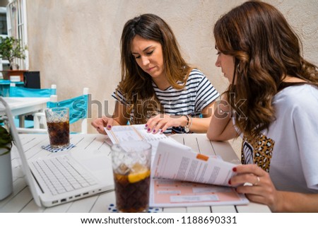Two girls in their twenties studying on a terrace with a drink and a computer. They are two white girls with brown hair. Terrace in light colours and white wood furniture.