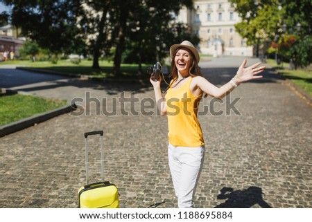 Overjoyed traveler tourist woman in yellow casual clothes with suitcase holding retro vintage photo camera spreading hands outdoor. Girl traveling abroad on weekend getaway. Tourism journey lifestyle