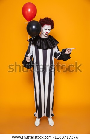 Young clown man 20s wearing black costume and halloween makeup grimacing and holding balloons isolated over yellow background