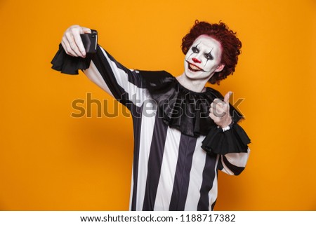 Happy clown man 20s wearing black costume and halloween makeup taking selfie photo on mobile phone isolated over yellow background