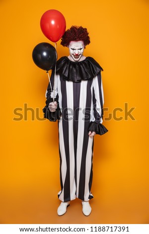 Ugly clown man 20s wearing black costume and halloween makeup grimacing and holding balloons isolated over yellow background