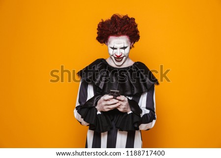 Dreadful clown man 20s wearing black costume and halloween makeup using smartphone isolated over yellow background