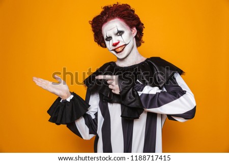 Horrific clown man 20s wearing black costume and halloween makeup holding copyspace at palm isolated over yellow background