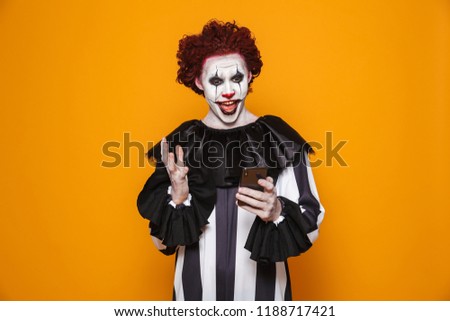 Young clown man 20s wearing black costume and halloween makeup holding smartphone isolated over yellow background