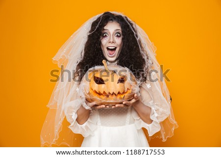 Image of dead bride zombie on halloween wearing wedding dress and scary makeup holding carved pumpkin isolated over yellow background