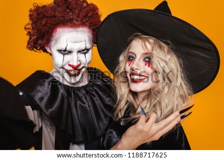 Photo of witch woman and clown man wearing black costume and halloween makeup taking selfie isolated over yellow background