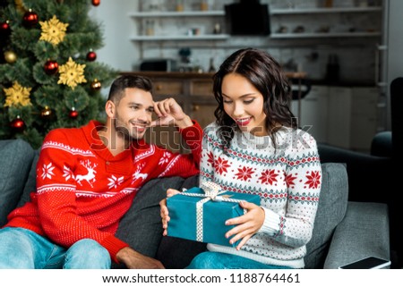 attractive young woman untying christmas present while her boyfriend sitting on sofa behind at home