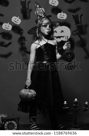 Girl with flirty face sends kiss on pink background with spooky decor. Little witch wearing black hat. Halloween party concept. Kid in spooky witches costume holds carved pumpkin and old gas lamp