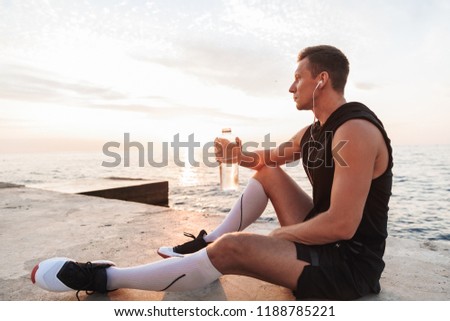 Picture of handsome strong young sportsman outdoors at the beach listening music with earphones drinking water.