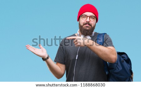 Young hipster man wearing red wool cap and backpack over isolated background amazed and smiling to the camera while presenting with hand and pointing with finger.