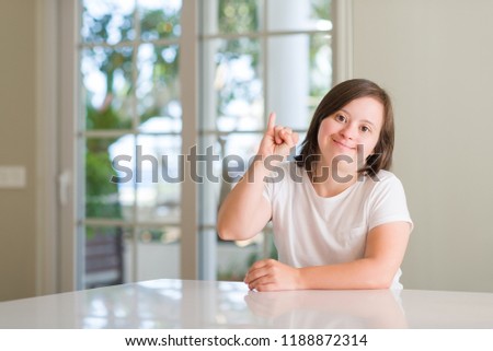 Down syndrome woman at home showing and pointing up with finger number one while smiling confident and happy.