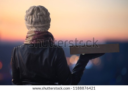 A girl in winter clothes with a box for pizza on the background of the evening city. 24 hours delivery. Concept 