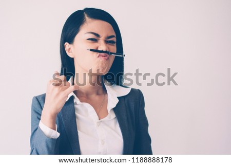 Closeup portrait of playful young pretty Asian business woman looking at camera and holding pen between lips and nose. Break concept. Isolated front view on grey background.