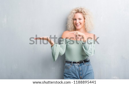 Young blonde woman with curly hair over grunge grey background amazed and smiling to the camera while presenting with hand and pointing with finger.