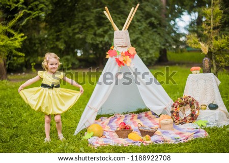 Little girl lying and playing in a tent, children's house wigwam in park Autumn portrait of cute curly girl. Harvest or Thanksgiving. autumn decor, party, picnic. Child in yellow dress with apples.