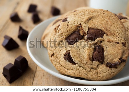 Close up picture of pile of chocolate cookies in white plate and a cup of milk and pieces of chocolate bar on wood table
