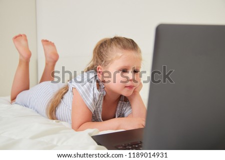 Cute little girl lying on bed at home, watching cartoons on laptop