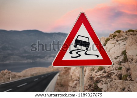 Photo of traffic sign (slippery road warning) with hills nad big mountains on background in Croatia. Black road with warning sign of slippery road and bad surface  at evening on sunset. 