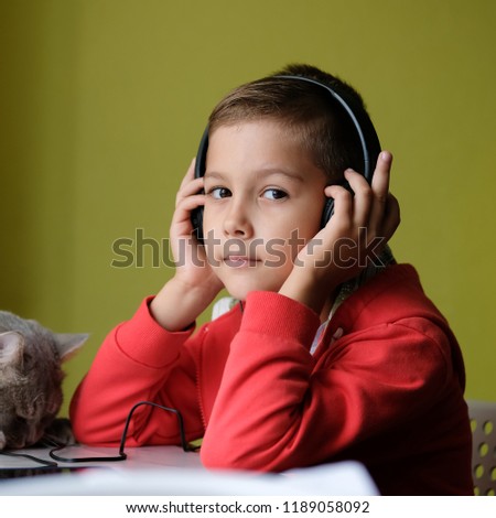 Young boy in his room listening to music with headphones. Child at the table watching a cartoon on the phone, and next is a cat.