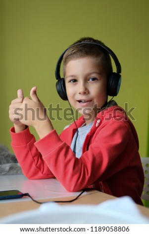 Young boy in his room playing phone with headphones. Child at the table listening to music and shows thumbs up. Kid puts a like.