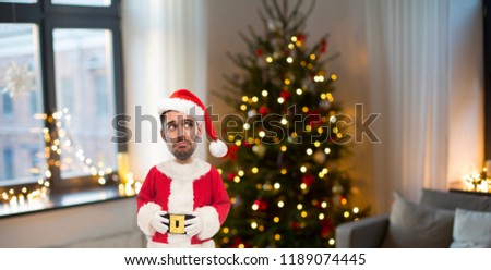 holidays, emotion, facial expressions and people concept - sad young man in santa clothes over christmas tree lights on background looking up (funny cartoon style character with big head)