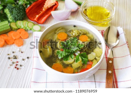 Light vegetable soup with ingredients of zucchini, broccoli, carrot, celery, pepper, onion, spices and a little olive oil on a white wooden background. Horizontal. Close-up.