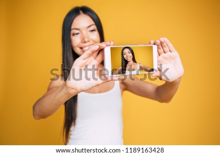 Happy cute smiling young asian brunette woman makes selfie photo of herself on smart phone isolated on yellow wall