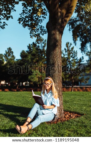 Portrait of cheerful beautiful coed girl, listening to music on headphones, sitting outdoors on green grass, wearing glasses, holding a book and reading it