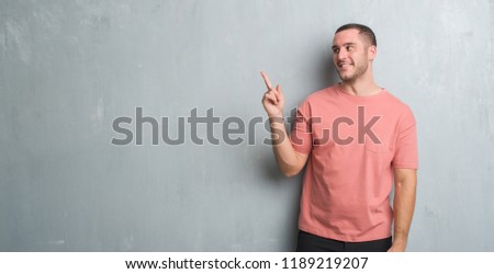 Young caucasian man over grey grunge wall with a big smile on face, pointing with hand and finger to the side looking at the camera.