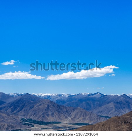 Desert hillside with bushes and tall barren mountains beyond under bright blue sky.