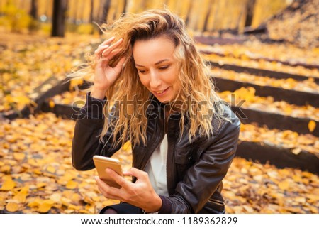 The girl is sitting on the stairs in the autumn forest. Brunette in a leather jacket with a phone in his hands, sms and internet surfing. Yellow leaf fall.