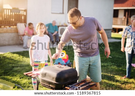 Mature man is taking care of the grill while his family is enjoying in backyard.
