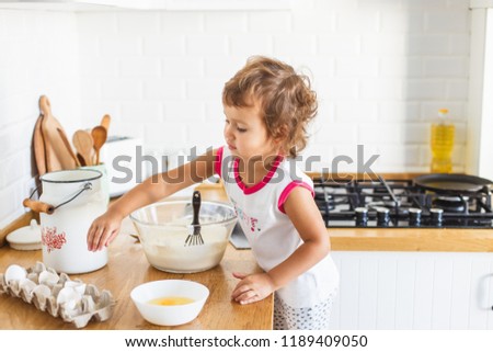 Little girl preparing dough for pancakes at the kitchen. Concept of food preparation, white kitchen on background. Casual lifestyle photo series in real life interior