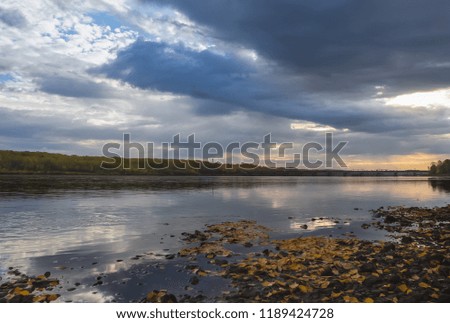 Panoramic picture of a calm river with blue waters, on which the yellow leaves lay, the sky is covered with clouds through which a ray of light tries to penetrate