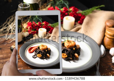 Photographing with tablet in hand.food concept with Black spaghetti. Black seafood pasta with shrimp in the white plate and red rose and grape wine. Mediterranean delicacy food. selective focus/still 