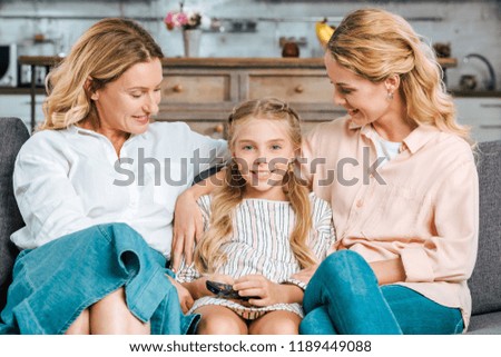child with mother and grandmother sitting on couch together with remote control at home