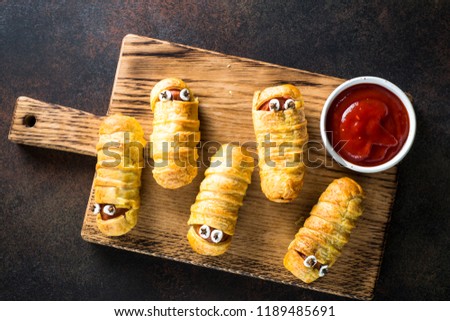 Halloween food. Scary sausage mummies in dough baked with funny eyes with ketchup on dark background. Funny decoration.  Top view.