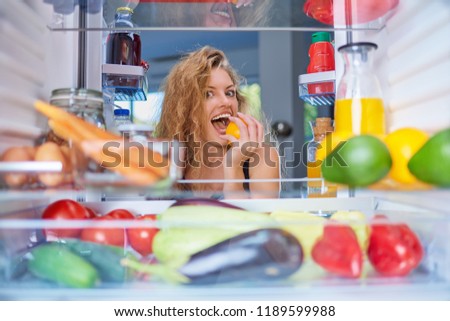 Woman biting fruit while standing in front of fridge full of groceries. Picture taken from the inside of fridge.