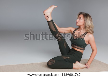 Young slim blond woman in sportswear sitting on mat holding leg up and stretching on gray background
