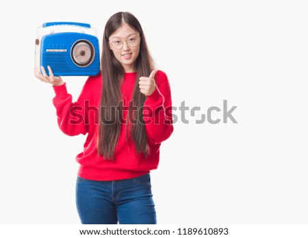 Young Chinese woman over isolated background holding vintage radio happy with big smile doing ok sign, thumb up with fingers, excellent sign