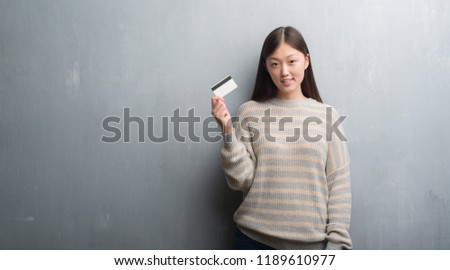 Young Chinese woman over grey wall holding credit card with a happy face standing and smiling with a confident smile showing teeth