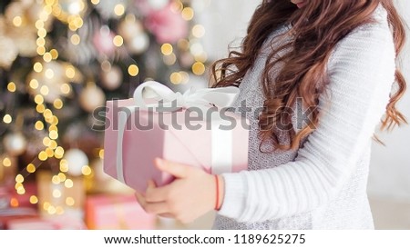 A little girl is holding a gift box in the hands of a Christmas tree.