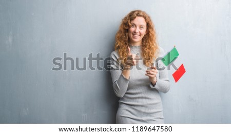 Young redhead woman over grey grunge wall holding flag of Italy happy with big smile doing ok sign, thumb up with fingers, excellent sign
