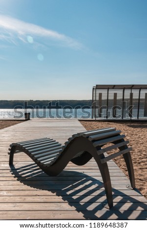 Beautiful wooden walkway and modern chairs at beach. Empty curved benches and trail among yellow sand. View of beach by river in sunny autumn day. Board way over sand. Bright sun makes deep shadows.