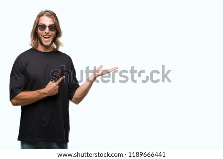 Young handsome man with long hair wearing sunglasses over isolated background amazed and smiling to the camera while presenting with hand and pointing with finger.