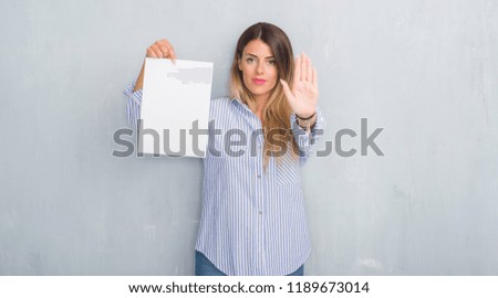 Young adult woman over grey grunge wall holding blank paper sheet with open hand doing stop sign with serious and confident expression, defense gesture