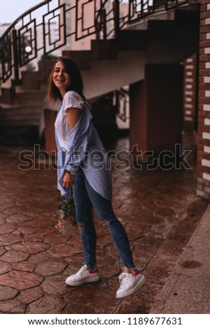 Half-turn photo of charming brunette woman with a dazzling smile holding flowers. Young girl walking in rainy weather carrying a bouquet of flowers. Dating outdoors, relationships