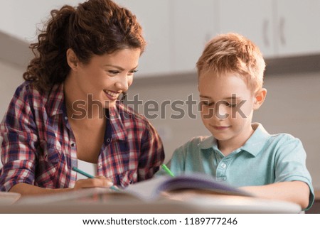 Young smiling mother watching her small boy coloring something and enjoying in their time at home.
