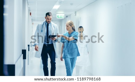 Female Surgeon and Doctor Walk Through Hospital Hallway, They Consult Digital Tablet Computer while Talking about Patient's Health. Modern Bright Hospital with Professional Staff. Royalty-Free Stock Photo #1189798168