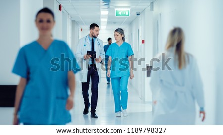 Surgeon and Female Doctor Walk Through Hospital Hallway, They Consult Digital Tablet Computer while Talking about Patient's Health. Modern Bright Hospital with Professional Staff. Royalty-Free Stock Photo #1189798327